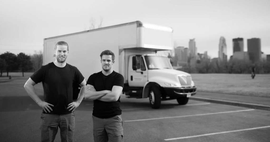 Black-and-white photo of two The Move Crew movers in black T-shirts standing in a parking lot in front of a white box truck, with a city skyline behind them.