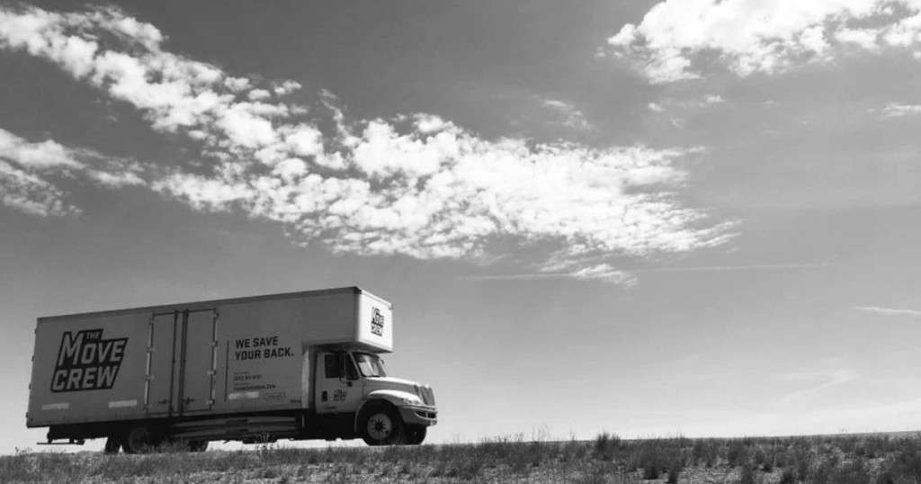 Black-and-white photo of a The Move Crew box truck driving across open grassland under a wide, cloudy sky